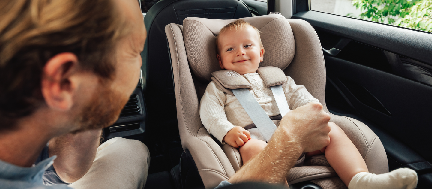 Een glimlachende baby zit veilig in een Britax R&ouml;mer Dualfix 5Z-autostoel in de kleur Teak, geplaatst op de achterbank van een auto. Een volwassene, van opzij gezien, houdt zachtjes het beentje van de baby vast terwijl hij of zij met hem communiceert.